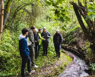 Five people are standing on a narrow, vegetated path next to the small Gembdenbach stream in the forest in Jena. They are looking at documents together and discussing.