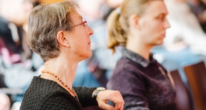 Dr. Wiebke Züghart sitzend, fokusiert von der Seite, konzentriert zuhörend im Plenum der Fachtagung. Im Hintergrund ist ein weiterer Teilnehmer schemenhaft zu sehen. 