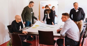 Um einen Tisch herum diskutieren vier Workshopteilnehmende. Die Moderatorin Frau Lachmann fässt die Diskussionspunkte auf dem Notizblatt auf dem Tisch zusammen unterstützt im Hintergrund von ihrer Kollegin.