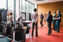 Several participants discuss in front of a moderation wall in a World Café format. The moderator notes down relevant points.