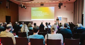A large audience seated in the plenary session, listening to a speaker at the podium.