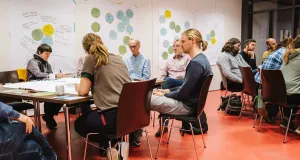 Participants are seated around two tables, writing on posters laid out on the tables.