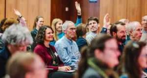 People in a plenary room listening attentively, smiling and sometimes raising their hands to ask questions or make contributions in a lively discussion during a forum.
