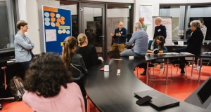 Eine Gruppe von Personen in einem Workshop-Raum, die in einem Diskussionskreis stehen oder sitzen. Im Hintergrund ist eine Pinnwand mit Notizen und Diagrammen zu sehen, während ein Teilnehmer eine Präsentation hält. Die Atmosphäre ist lebendig und gemeinschaftlich.