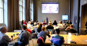 Zahlreiche Menschen sitzen im Plenum und lauschen dem Onlinevortrag, der auf der Leinwand im Hintergrund zu sehen ist