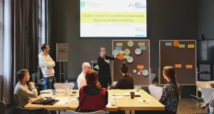 Einige Menschen sitzen um einen Tisch. Die Moderatorin steht hinter ihnen. Im Hintergrund stehen zwei Pinnwände, auf denen bunte Karteikarten mit Notizen angebracht sind.