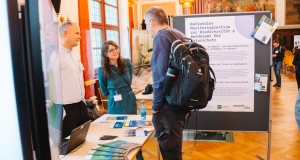 Mitarbeitende des BfN und des Monitoringzentrums informieren die Gäste zu aktuellen Aktivitäten und beantworten Fragen. Im Hintergrund ist ein Poster des Monitoringzentrum zu sehen.