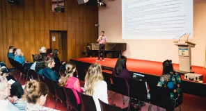 Plenumssituation, auf der Bühne steht der Moderator, im Vordergrund sind die Teilnehmenden von hinten zu sehen