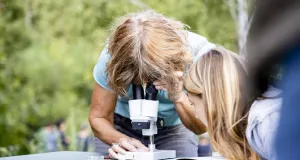 Eine Person betrachtet eine Probe durch ein Mikroskop im Freien, während eine zweite Person rechts davon neugierig zuschaut.