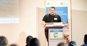 Thomas Hörren, Vorsitzender Entomologischer Verein Krefeld steht am Rednerpult und hält seinen Impulsvortrag auf dem 2. Forum des NMZB. Im Hintergrund steht ein Roll-up des Forums, im Vordergrund sitzen einige Teilnehmende, diese sind von Hinten fotographiert.