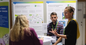 Zwei Frauen und ein Mann stehen um einen Tisch und unterhalten sich. Im Hintergrund sind zwei Poster am Infostand des Monitoringzentrum zu sehen