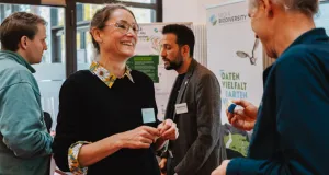 Mehrere Personen stehen im Austausch vor einem Infostand von NFDI4Biodiversity. Eine Kollegin des Monitoringzentrums tauscht sich mit einem Kollegen von NFDI4Biodiversity aus, im Hintergrund sind Poster und Roll-up mit dem Slogan „Daten. Vielfalt. Artenvielfalt.“ zu sehen. Der steht im Fokus.