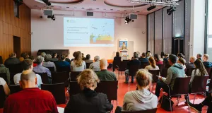Im Hintergrund steht eine Frau am Podium und hält einen Vortrag. Im Vordergrund sitzt das Publikum in zahlreichen Stuhlreihen.