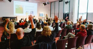 Im Hintergrund steht eine Frau am Podium und befragt das Publikum. Im Vordergrund sitzt das Publikum in zahlreichen Stuhlreihen. Einige Personen heben in einer Abstimmung die Hand.