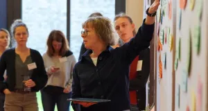 Eine Frau zeigt auf verschiedene Zettel an einer Pinnwand, im unscharfen Hintergrund stehen weitere Personen 