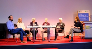 Three women and three men sitting in a semicircle on a stage, engaging in a discussion.