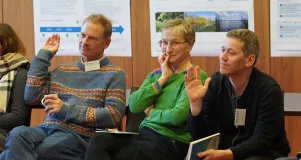 Eine Frau und zwei Männer sitzen nebeneinander, die Männer heben jeweils ihr rechte Hand für eine Abstimmung im Workshop