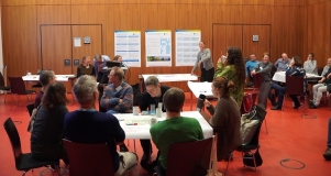 Es sitzen acht Menschen um einen Tisch und besprechen sich, im Hintergrund sitzt eine weitere Gruppe. 