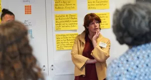 Eine Frau stet vor einer Pinnwand. Um sie herum sind große Zettel mit Inhalten aus dem Workshop angebracht