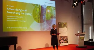 Eine Frau steht alleine auf einer Bühne. Hinter ihr ist eine Präsentation auf der Wand sichtbar. a 