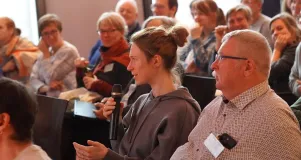 Eine Frau sitz im Publikum und hält Mikrofon in der Hand, um eine Frage zu stellen