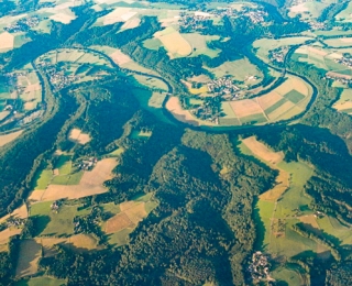 Luftbild einer strukturreichen Landschaft mit Äckern, Grünland und Forst durch die ein Fluss mäandriert.