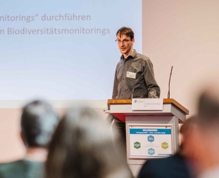 Im Hintergrund ist auf Dr. David Eichenberg am Rederpult fokussiert, welcher referiert. Im Vordergrund sind einige Teilnehmende im Plenum von hinten zu sehen.