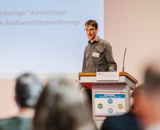 Im Hintergrund ist auf Dr. David Eichenberg am Rederpult fokussiert, welcher referiert. Im Vordergrund sind einige Teilnehmende im Plenum von hinten zu sehen. 