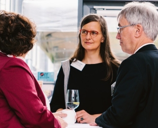 In conversation at a high table, Sabine Riewenherm (left), President of BfN, can be seen from the back on the left, wearing a red jacket; in the middle, Dr Petra Dieker, the new director of the Monitoring Centre, in a black dress, with long light brown straight hair and red glasses; to her right, from the side, Dr Andreas Krüß in a black jacket, with mottled grey hair and glasses.