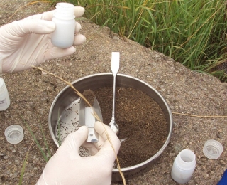 In the middle of the photo there is a soil sample in an open round metal container. Two hands with white laboratory gloves can be seen dividing the sample into subsamples. These are filled into small white containers.