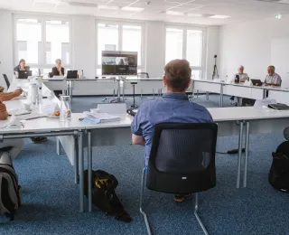 Several people sit together at tables in a rectangular shape. In the middle there is a screen through which other participants are joining online.