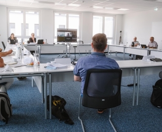 Several people sit together at tables in a rectangular shape. In the middle there is a screen through which other participants are joining online.