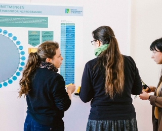 Drei Kolleginnen des Monitoringzentrums von der Seite und von hinten zu sehen, stehen vor einem Poster zur Netzwerkanalyse.
