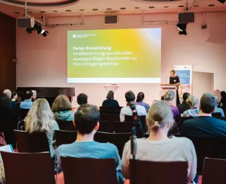 Zahlreiche Menschen sitzen im Plenum und lauschen der Referentin am Podium