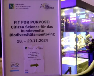 Im Vordergrund steht das Hinweisschild an der Treppe zum Seminarraum des Workshops, im Hintergrund ist verschwommen ein Dinosaurierschädel in einer Vitirine zu sehen