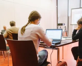 Im Vordergrund werden Mitschriften am Laptop getätigt. Im Hintergrund sitzen Teilnehmde und diskutieren. 