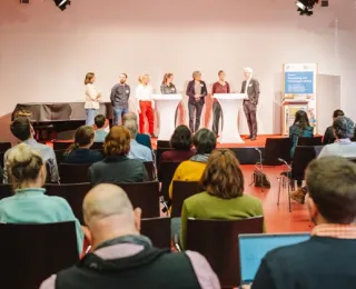 Mehrere Mitglieder des Grundsatzfachgremiums stehen auf einer Bühne an Stehtischen und geben Kurzimpulse in einer Blitzlichtrunde als inhaltlicher Auftakt des 3. Forums des Monitoringzentrums. Das Publikum ist von hinten zum Teil zu sehen. 