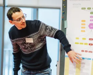 Ein Mann mit kurzen braunen Haaren, Brille und gemustertem Pullover steht in einer Workshopsituation neben einer Posterwand und zeigt mit der Hand auf eine detaillierte Diagramm- und Textübersicht. Im Hintergrund ist ein Fenster.